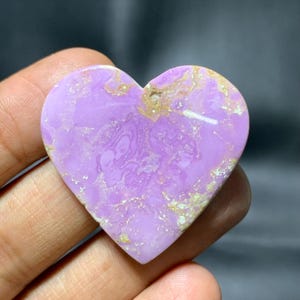 May include: A heart-shaped gemstone in a light purple shade, featuring detailed swirling patterns. The stone has gold and tan accents, contrasting with its smooth, polished surface. The gemstone is held in a hand.