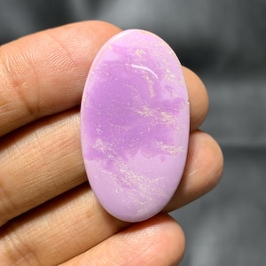 May include: A polished, oval gemstone in a soft lavender colour. The stone has a smooth surface with subtle, swirling white inclusions, creating a marbled effect. The gemstone is held in a hand, showcasing its size and colour.