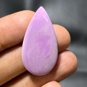 May include: A polished, teardrop-shaped gemstone in a pale lavender colour. The stone has a smooth, glossy surface with subtle, lighter-coloured veining. The gemstone is held in a hand, showing its size and shape. The overall aesthetic is elegant and natural.