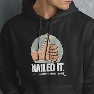 Op de afbeelding: Zwarte hoodie met een afbeelding van een hand die een duim omhoog steekt in een lichtblauwe cirkel. De tekst "NAILED IT." staat onder de afbeelding, met "prompt: human hand" eronder. De hoodie heeft een voorzak en een capuchon.
