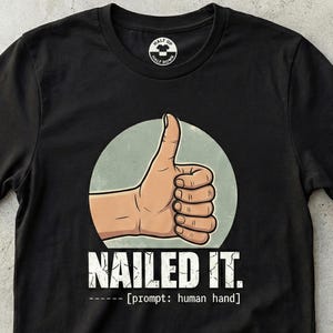 Op de afbeelding: Zwart t-shirt met een cartoon hand die een duim omhoog geeft in een lichtgroene cirkel. De woorden "NAILED IT." staan eronder in witte, vette letters. De tekst "[prompt: human hand]" staat eronder.