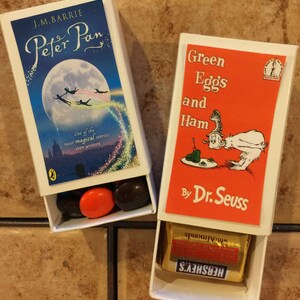 May include: Two matchbox-style boxes with book covers. The first box has a blue cover with a moon and stars, and the text "J.M. Barrie Peter Pan". The second box has a red cover with the text "Green Eggs and Ham by Dr. Seuss". Both boxes have candy inside.