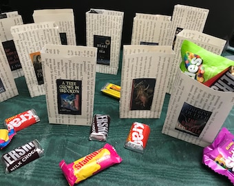 Può includere: Un tavolo con una tovaglia verde e diversi piccoli sacchetti di carta decorati con copertine di libri. I sacchetti contengono barrette di cioccolato e una busta di Skittles. I titoli dei libri includono "A Tree Grows in Brooklyn", "The Outer Triangle", "Greys", "Heart of the Sea" e "The Book Thief".