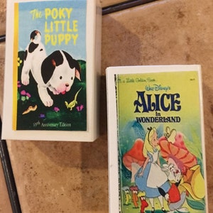 May include: Two miniature book covers. The first cover is blue and yellow with a black and white puppy on it. The title is "The Poky Little Puppy" and the text "65th Anniversary Edition" is at the bottom. The second cover is yellow and green with a picture of Alice from Alice in Wonderland. The title is "Walt Disney's Alice in Wonderland".