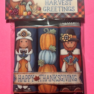 May include: A clear plastic package containing three cylindrical items with Thanksgiving-themed illustrations. The top label reads HARVEST GREETINGS, and a banner across the center reads HAPPY THANKSGIVING with a maple leaf graphic.