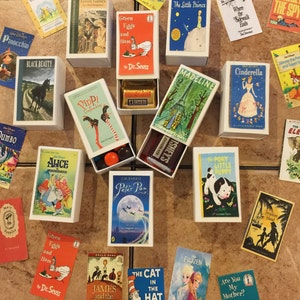 May include: A collection of children's book matchboxes, including classics like 'The Little Prince', 'Madeline', 'Where the Sidewalk Ends', 'Alice in Wonderland', 'Peter Pan', 'Green Eggs and Ham', 'The Cat in the Hat', 'James and the Giant Peach', 'Frozen', 'Are You My Mother?', 'The Portly Little Puppy', 'Treasure Island', 'Winnie the Pooh', 'Black Beauty', 'Pinocchio', 'Dumbo', 'Pippi Longstocking', 'Anne of Green Gables', and 'Curious George'.
