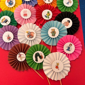 May include: A collection of colorful paper rosette cupcake toppers. Each rosette is a different color and features a circular sticker with a cartoon character. The toppers are arranged on a red surface.