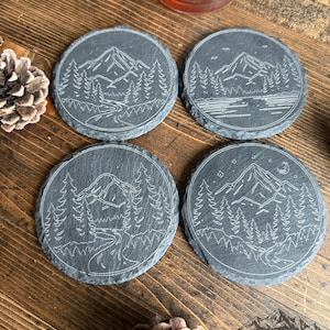 May include: Four round slate coasters with etched mountain and forest designs. Each coaster features a unique landscape scene with trees, mountains, and a river. The coasters are dark gray and rest on a wooden surface.