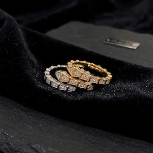 May include: Three snake-shaped rings, one gold and two silver, are displayed on a black velvet surface. The gold ring is encrusted with small, clear gemstones. The silver rings are also adorned with clear gemstones. The rings are in a coiled position.