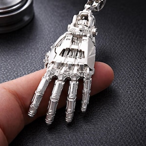 May include: A silver-colored, articulated robotic hand pendant. The pendant features detailed mechanical components, including fingers, knuckles, and a wrist joint. The design is highly detailed and realistic, with a metallic finish. The pendant is likely made of metal.