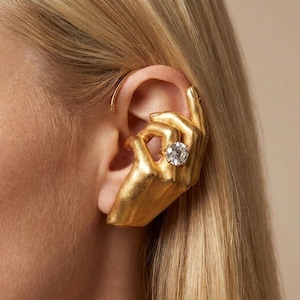 Puede incluir: Un pendiente de oreja dorado con forma de mano, sosteniendo un gran anillo de piedra preciosa transparente y de corte redondo. El pendiente abraza la oreja, con los dedos de la mano delicadamente colocados. La joya se encuentra sobre un fondo neutro.