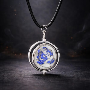May include: A silver-toned pendant necklace featuring a circular design with a blue and white floral pattern. The pendant hangs from a black cord. The necklace is displayed against a dark background.