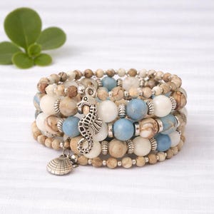 May include: Stack of beaded bracelets with a beach theme. The bracelets feature a mix of light blue, cream, and brown beads, accented with silver-toned spacers. A seahorse and seashell charm dangle from the bracelets.