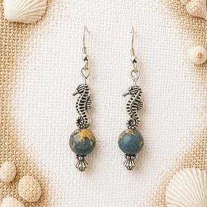 May include: A pair of silver seahorse earrings with blue and gold beaded accents. The earrings are displayed on a white surface with seashells in the background, evoking a coastal theme.
