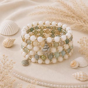 May include: A stack of beaded bracelets with white, green, and tan beads, accented with gold-colored spacers. Two seashell charms dangle from the bracelets. Seashells and dried plants surround the bracelets, creating a beachy theme.