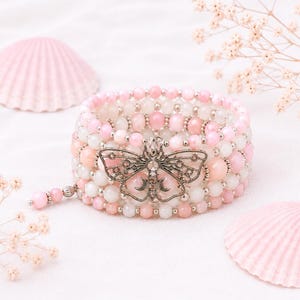 May include: A multi-strand beaded bracelet featuring alternating light pink and white spherical beads. A silver-toned metal moth charm with intricate wing patterns and a crescent moon detail is attached to the center of the bracelet.