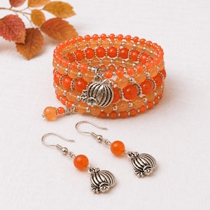 May include: A multi-strand wrap bracelet and a pair of matching drop earrings featuring orange beads and silver-toned pumpkin charms. The bracelet is constructed with several rows of round orange beads and small silver spacers, while the earrings consist of a single orange bead and a silver pumpkin charm on a hook wire.