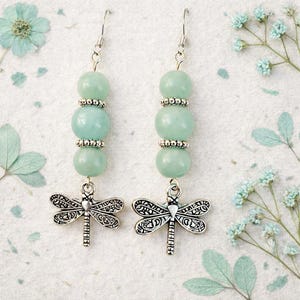 May include: A pair of dangle earrings featuring three light green beads separated by silver-colored spacers. Each earring has a detailed silver dragonfly charm suspended below the beads. The earrings hang from silver-colored ear wires.