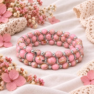 May include: A multi-strand stretch bracelet featuring round pink marbled beads interspersed with small, shiny silver-toned metal spacer beads. The beads exhibit a smooth, polished finish with subtle variations in pink and white tones throughout.