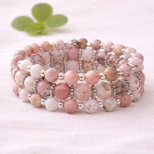 May include: Three-strand bracelet featuring round, pink and white gemstone beads. The beads are interspersed with small silver-colored beads. The bracelet is displayed on a white surface, with a green leaf in the background.