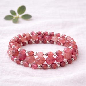 May include: A multi-strand bracelet featuring pink and red gemstone beads. The bracelet has three rows of beads, with a mix of round and irregular shapes, separated by small silver-colored beads. The bracelet is displayed on a white surface.