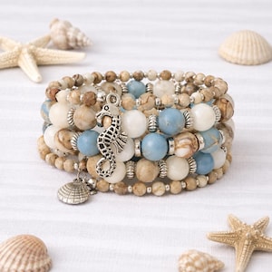 May include: A multi-strand beaded bracelet featuring a mix of round tan, brown, and light blue beads accented with silver-toned spacers. The bracelet includes a silver-toned seahorse charm and a small silver-toned seashell charm hanging from it.