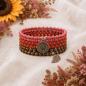 May include: A multi-strand beaded bracelet featuring rows of pink, red, and brown spherical beads separated by small silver-toned spacers. The bracelet includes a round silver-toned sunflower charm and a silver-toned leaf-shaped charm.