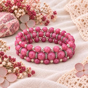 May include: A multi-strand stretch bracelet featuring round, polished magenta-pink stone beads interspersed with small, reflective silver-toned metal spacer beads arranged in a repeating pattern around the entire circumference.
