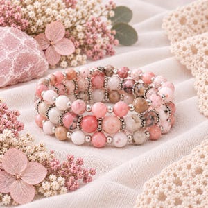 May include: A multi-strand beaded bracelet featuring round polished stones in shades of pink, white, and tan. The beads are interspersed with small, textured silver-toned metal spacers, creating a layered appearance with a consistent circular design.