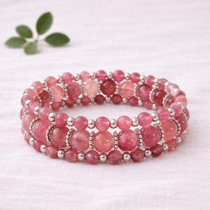 May include: A multi-strand bracelet featuring pink and red gemstone beads. The bracelet has three rows of beads, with larger, round beads in the center row and smaller beads on the top and bottom rows. Silver-colored metal accents separate the rows.