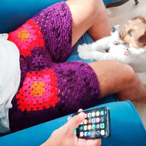 May include: Crochet shorts featuring a dark purple base with vibrant granny square motifs in shades of bright pink and orange. The shorts have a textured, open-stitch pattern throughout. The image includes the text UknitMe2 and PDF.