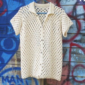 May include: A cream-coloured, short-sleeved crochet shirt featuring a collared neckline and a front button closure. The garment is constructed with an open, repeating square stitch pattern. The text UknitMe2 and PDF are visible at the top.