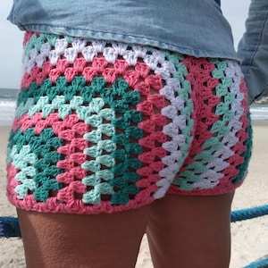 May include: Crochet shorts featuring a granny square pattern in alternating rows of white, light mint green, dark teal, and coral pink yarn. The shorts have a visible open-stitch texture and are worn with a blue denim shirt. Text reads UknitMe2 and PDF.