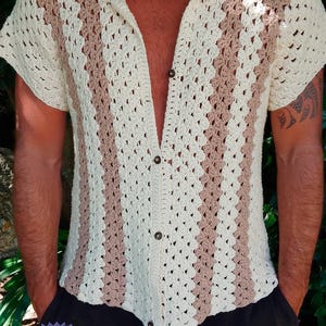 Può includere: Una camicia all'uncinetto a maniche corte con strisce verticali color crema e marrone chiaro. Il capo presenta un colletto, un'abbottonatura frontale con quattro bottoni scuri e una trama a maglia aperta. Il testo PDF e UknitMe2 è visibile in basso.