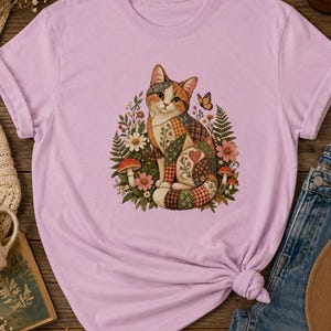 Può includere: Una t-shirt color viola chiaro con una grafica centrale raffigurante un gatto dal manto in stile patchwork seduto tra fiori colorati, funghi e una piccola farfalla. Il gatto presenta un motivo a forma di cuore sul fianco ed è circondato da foglie.