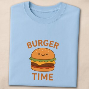 Könnte beinhalten: Hellblaues Rundhals-T-Shirt mit einem Cartoon-Burger-Design. Der Burger hat ein lächelndes Gesicht und besteht aus Brötchen, Salat, Käse und Tomate. Die Worte "BURGER TIME" sind in Orange über und unter dem Burger aufgedruckt.