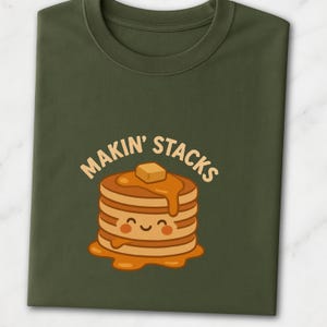Puede incluir: Camiseta verde oliva con una pila de panqueques de dibujos animados, mantequilla y jarabe, con la frase "MAKIN' STACKS". Los panqueques tienen una cara sonriente. Una divertida camiseta gráfica con temática de comida.
