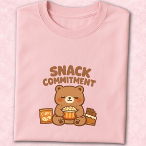 Może przedstawiać: Różowa koszulka z okrągłym dekoltem z kreskówkowym misiem trzymającym popcorn, z paczką chipsów i batonem czekoladowym. Tekst "SNACK COMMITMENT" znajduje się nad misiem w brązowych literach.
