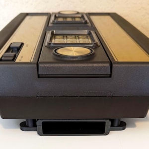 May include: A vintage black and gold video game console. The console has a rectangular shape with a gold panel on top, featuring buttons and a joystick. The cartridge slot is visible at the bottom. A classic gaming device.