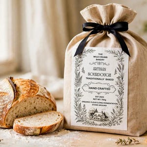 May include: A round loaf of sourdough bread with a sliced piece next to a beige linen drawstring bag tied with a black ribbon. The bag features a white label with the text: THE WILD CRUMB BAKERY, ARTISAN SOURDOUGH, TRADITIONALLY BAKED, HAND CRAFTED, EST 2023, NET WT: 750g, ORGANIC FLOUR LONG FERMENTATION, STONE GROUND.