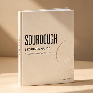 May include: A hardcover book with a cream-colored cover titled SOURDOUGH BEGINNER GUIDE A Modern Guide to Your First Loaf in black text. The cover features a subtle embossed circular line design and the brand name AASHZAM at the bottom.