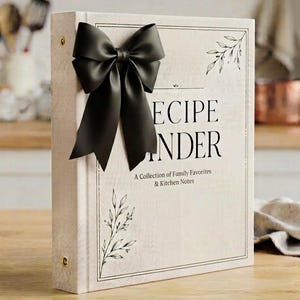 May include: A cream-colored recipe binder featuring a large black satin bow on the front cover. The text reads RECIPE BINDER A Collection of Family Favorites & Kitchen Notes. The cover includes black line art illustrations of leafy branches.