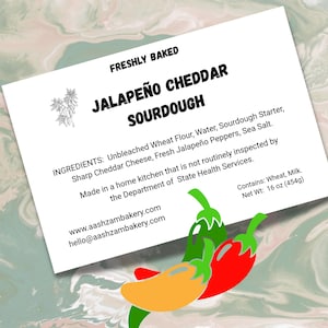 May include: A white label with text reading "Freshly Baked Jalapeño Cheddar Sourdough." Ingredients include wheat flour, cheddar cheese, and jalapeño peppers. The label also features a drawing of chili peppers and states the net weight is 16 oz (454g).