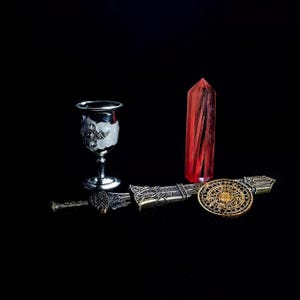 Puede incluir: Un surtido de artículos de fantasía sobre un fondo negro. Incluye un cáliz plateado con detalles ornamentados, un obelisco de cristal rojo, una espada decorativa con vaina y un emblema circular dorado.