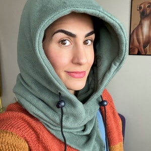 Puede incluir: Un cuello de cuello verde de forro polar con capucha, cordones negros y cierres de bot&oacute;n negros. El cuello de cuello est&aacute; siendo usado por una persona con cabello casta&ntilde;o y ojos marrones.