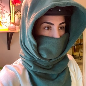 Puede incluir: Un calentador de cuello con capucha de color verde oscuro de forro polar con una cubierta para la cara. El calentador de cuello est&aacute; doblado hacia abajo para mostrar el forro polar.