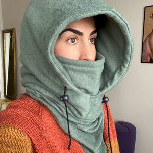 Puede incluir: Una balaclava de forro polar verde con un cord&oacute;n de ajuste y botones negros. La balaclava se tira sobre la cabeza y cubre la cara, dejando solo los ojos visibles.