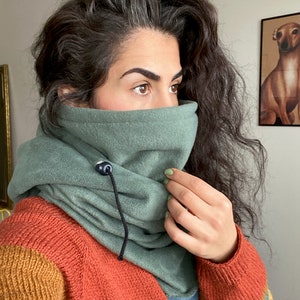 Puede incluir: Un cuello de lana verde con un cord&oacute;n de ajuste negro y un bot&oacute;n negro. El cuello de lana est&aacute; siendo usado por una persona con cabello largo y oscuro.