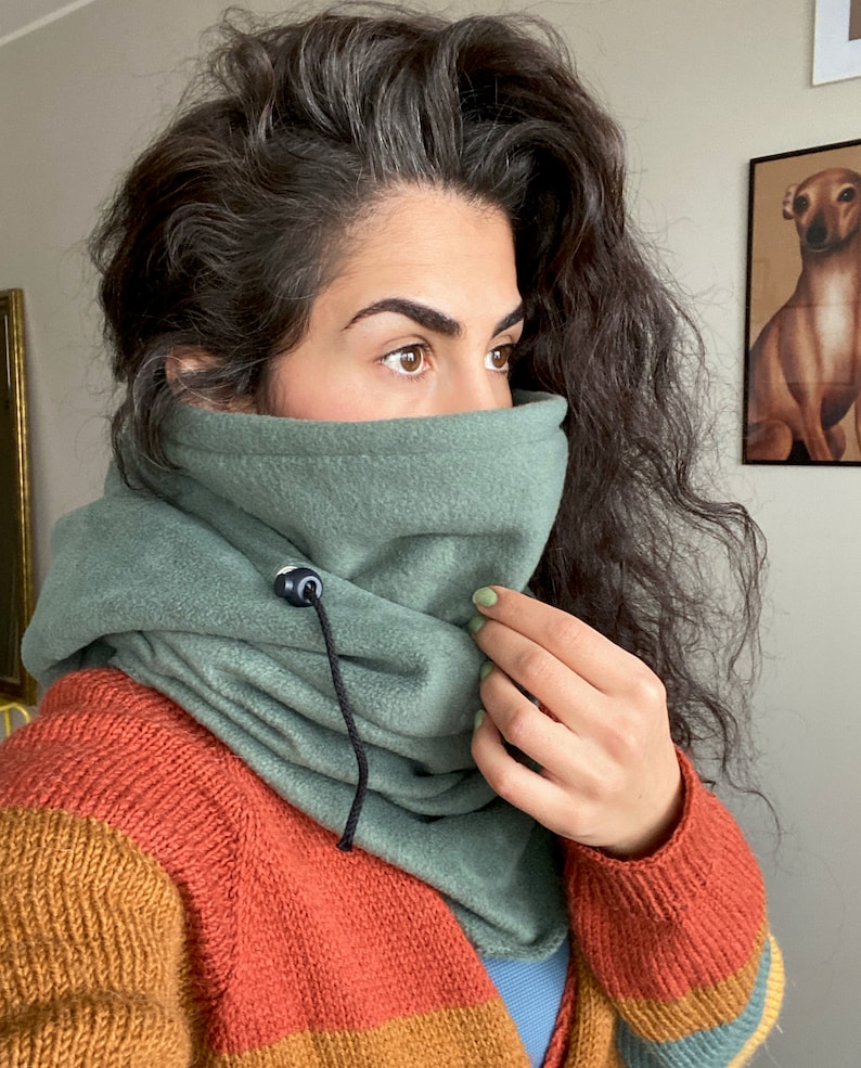 Puede incluir: Un cuello de lana verde con un cord&oacute;n de ajuste negro y un bot&oacute;n negro. El cuello de lana est&aacute; siendo usado por una persona con cabello largo y oscuro.