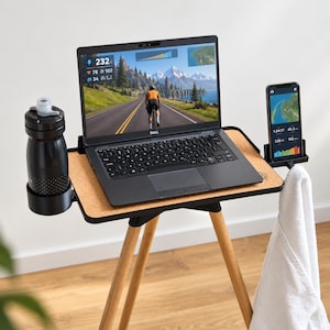 Könnte beinhalten: Ein tragbarer Trainingsschreibtisch mit Korkoberfläche, ausgestattet mit einem schwarzen Dell-Laptop, der eine Radsimulation anzeigt, einer schwarzen Trinkflasche im Seitenhalter, einem Smartphone und einem weißen Handtuch am Rahmen.